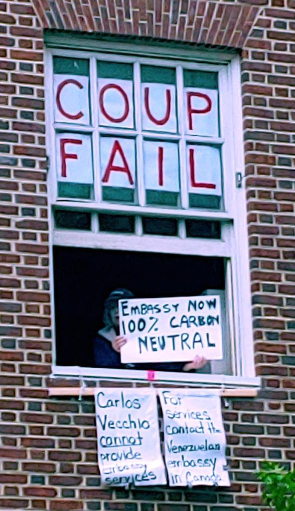 Photo of an embassy protector in a darkened window, holding a sign that reads "Embassy now 100% Carbon Neutral." Around the figure are other signs that say: "COUP FAIL," "Carlos Veccio cannot provide embassy services," "For services contact the Venezuelan embassy in Canada."