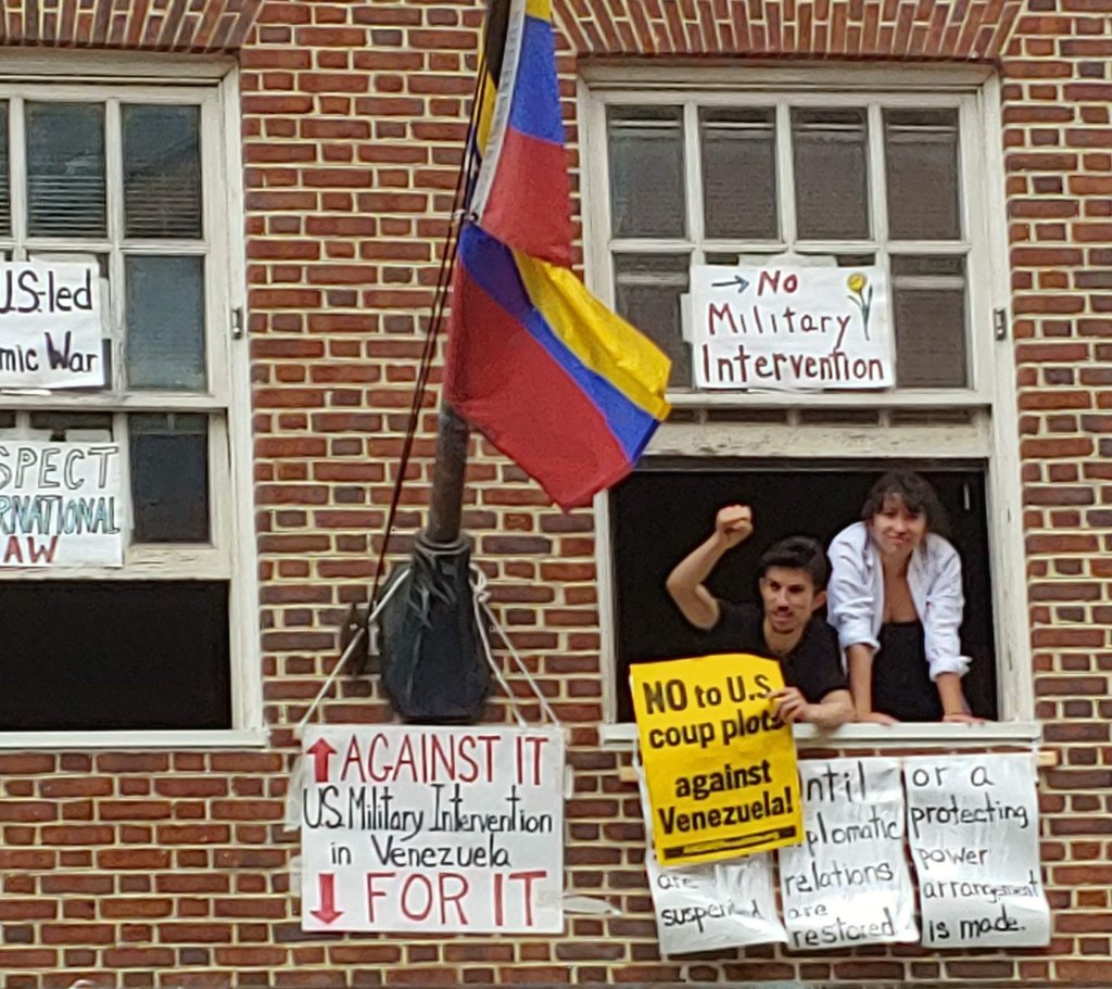 Photograph of two Embassy Protectors, a man and a woman, in an upstairs window of the Venezuelan Embassy. The man is raising his fist in a gesture of defiance. Over their heads is a sign that reads "No Military Intervention."