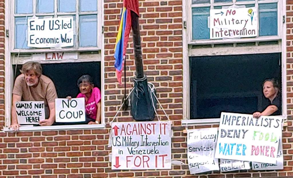 A close-up photograph of three Embassy Protectors in their upper-floor windows. They hold signs which read: "Guaido not welcome here," "No coup," "Imperialists deny food, water, power."