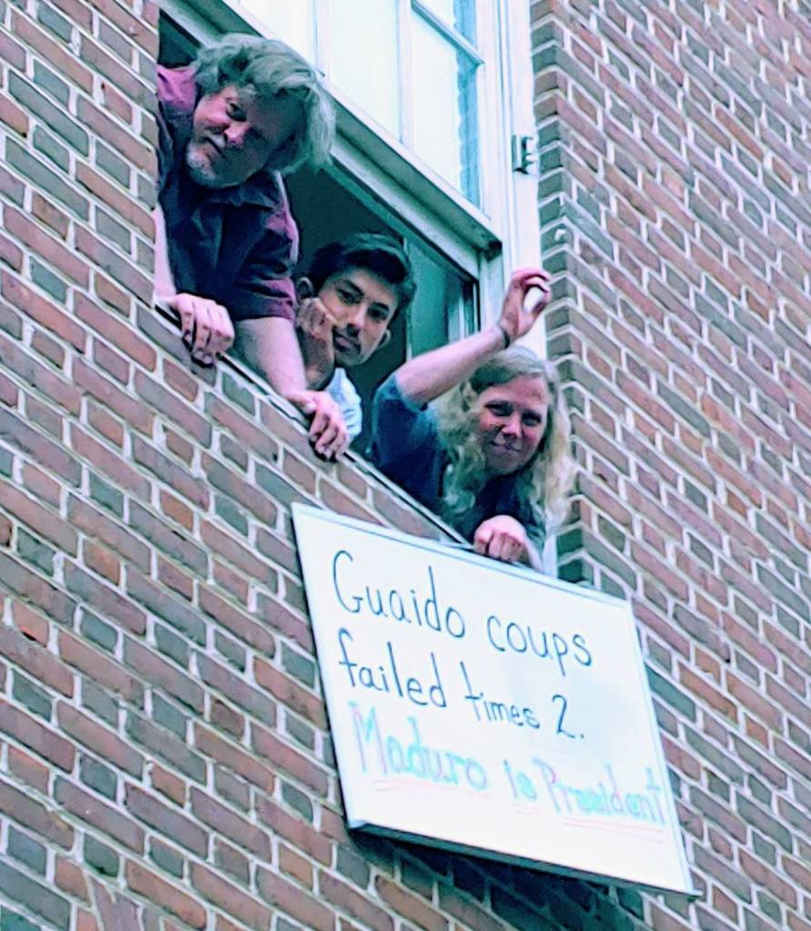 Photograph of three Embassy Protectors, two men and a woman, leaning out of a window and holding a sign which reads "Guaido coups failed times 2. Maduro is President."