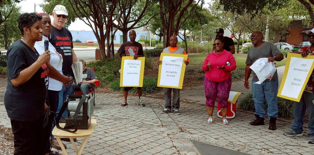 Photo of a Black woman speaking to nine people attending an outdoor press conference. The group is majority-Black. Four hold signs: "Let the Truth be Told, Justice Now"; "Free Jermaine Doss, Wrongfully Accused"; "Free Jermaine Doss, Wrongfully Convicted."