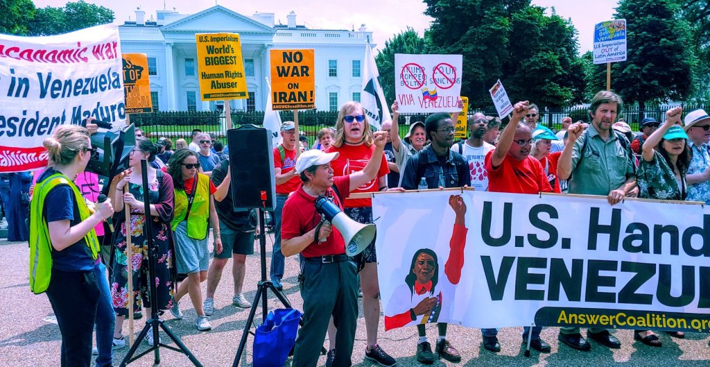 Photograph of a large, multi-racial march, with many banners and signs, standing in front of the White House. Various signs read: "U.S. Imperialism: World's biggest human rights abuser"; "No war on Iran"; "Viva Venezuela"; "U.S. Imperialism Out of Latin America". 