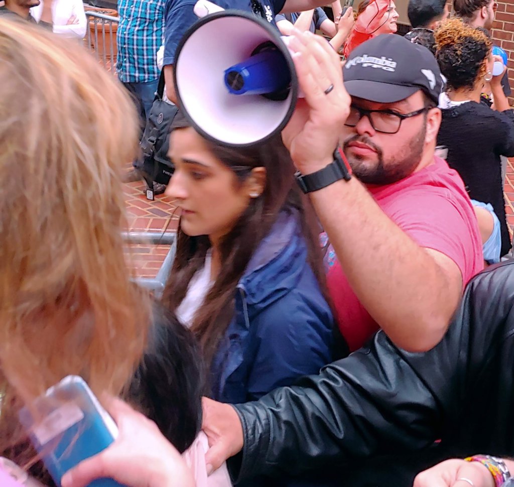Photograph of a man holding a bullhorn above his head, amidst a crowded scene which appears to be a conflict. 
