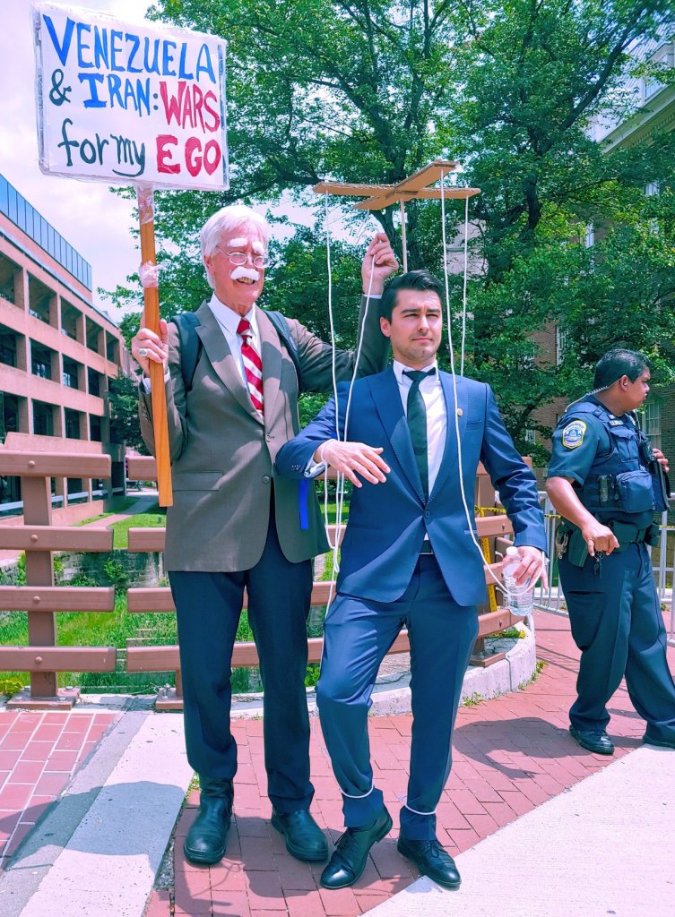 Photograph of two men: one is dressed as a satirical depiction of U.S. National Security Advisor John Bolton and holds up a sign which reads, "Venezuela and Iran: Wars for my Ego". In his other hand he holds an apparatus to control a marionette, which is connected to the other man with strings. The other man, who is posing as though he is a puppet, is dressed like Carlos Vecchio. 