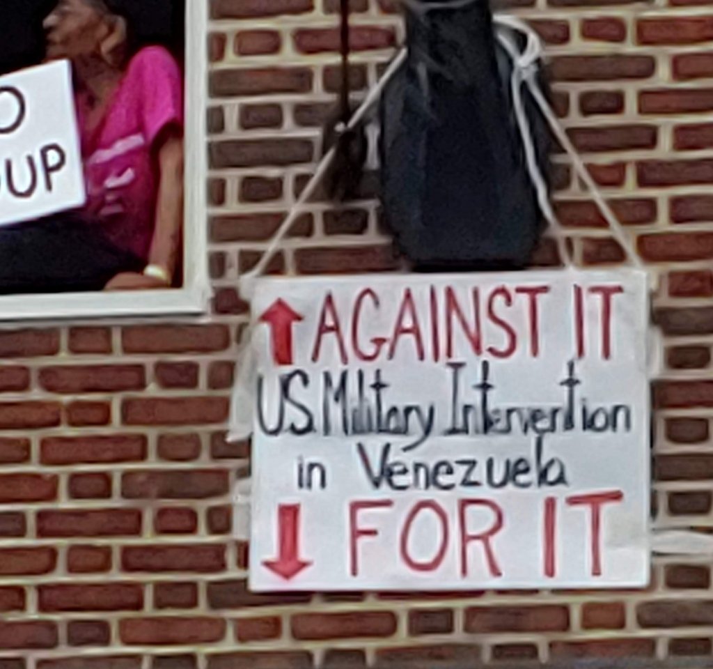 Photograph of a sign hung on the Venezuelan embassy. It reads: "U.S. Military Intervention In Venezuela -- Against It, For It". There is an arrow pointing up to indicate that those in the upper windows are against it, and an arrow pointing down to indicate that the crowd at street level is for it. 