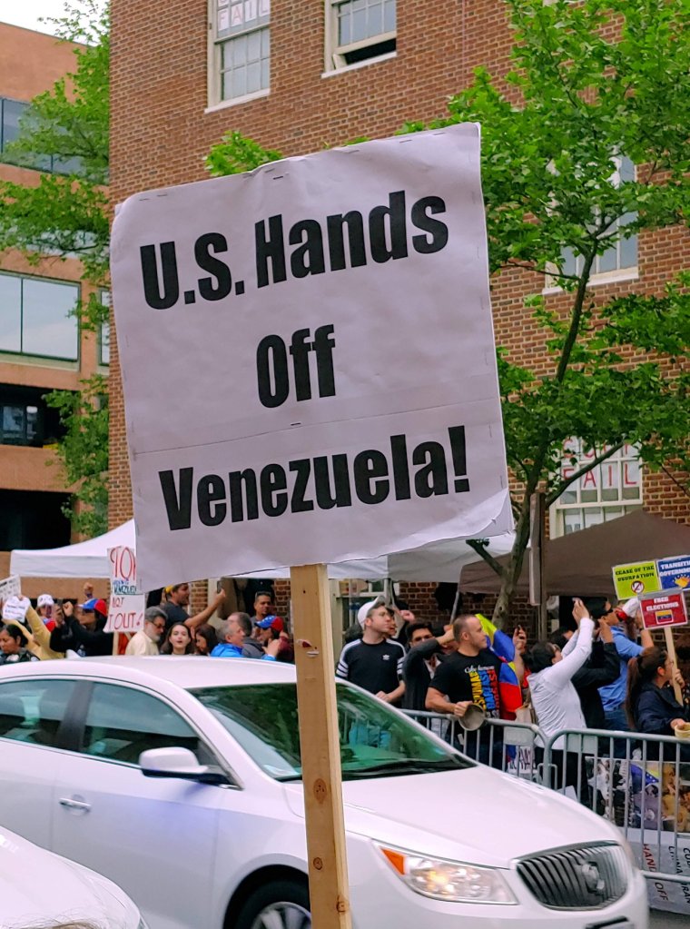Photograph of a sign which reads "U.S. Hands Off Venezuela!" in the foreground, with a view of a large crowd clustered around a brick building behind it. 