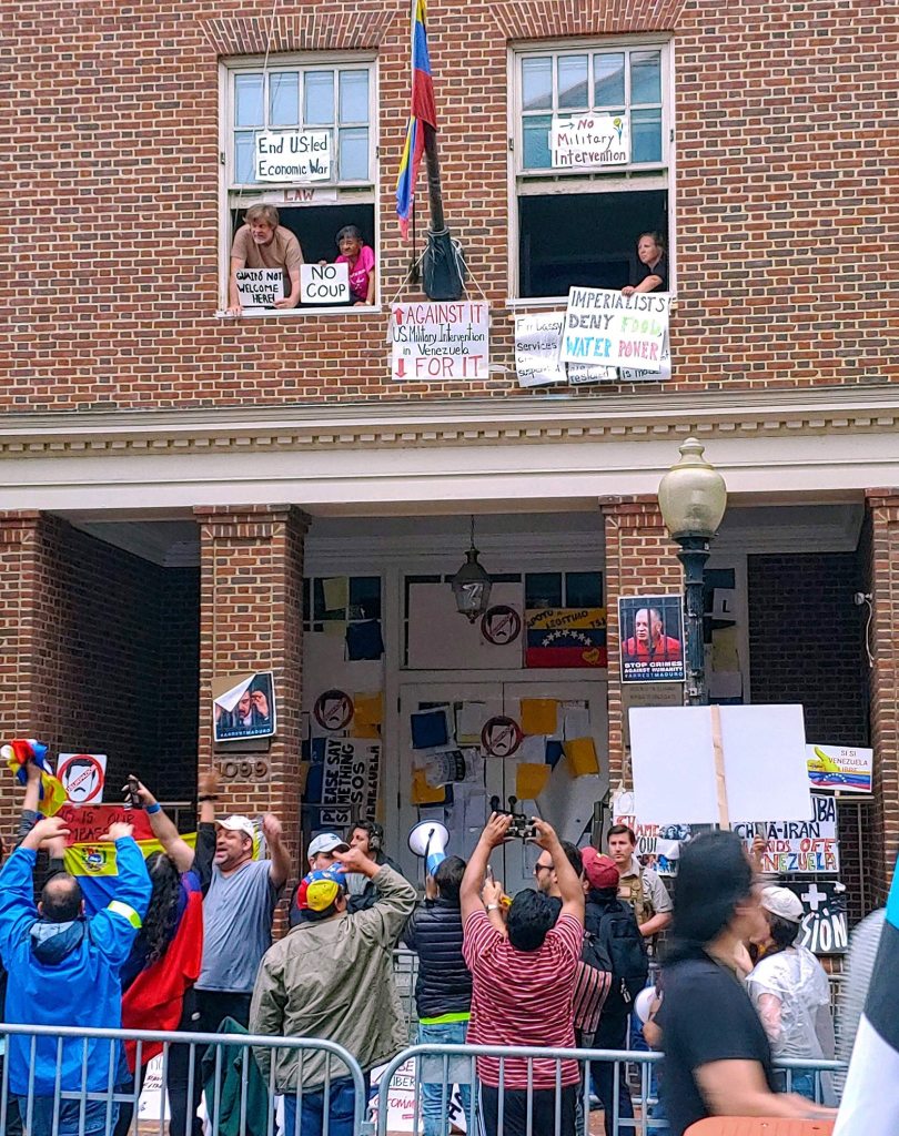 Photograph of the Venezuelan Embassy with a large crowd of coup supporters gathered around it as occupiers of the building look on from second-story windows festooned in anti-coup signs. 