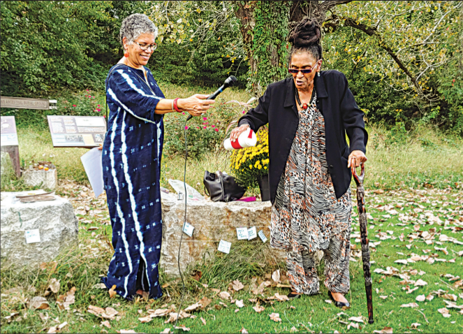 Queen Nzinga, right, speaks and pours water from a container while Ana Edwards, left, holds a microphone for her. They stand outdoors among fallen leaves. 