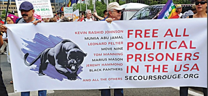 Photograph of people holding a banner emblazoned with the phrase "Free all political prisoners in the USA," a short list of such prisoners, and an image of a black panther. 