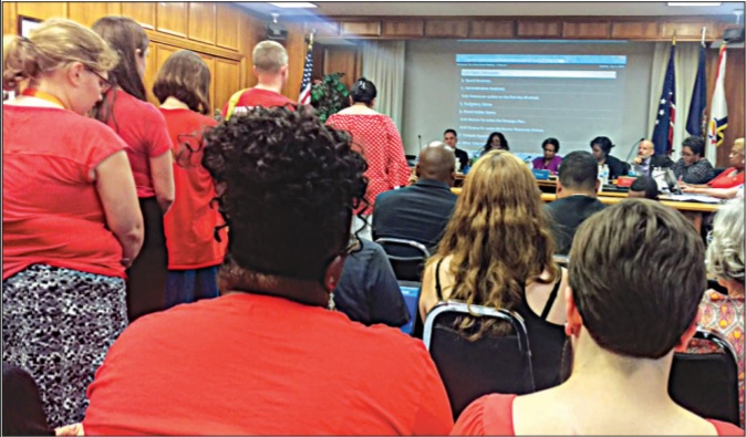 Photograph of a crowded school board meeting, with many audience members wearing red. 