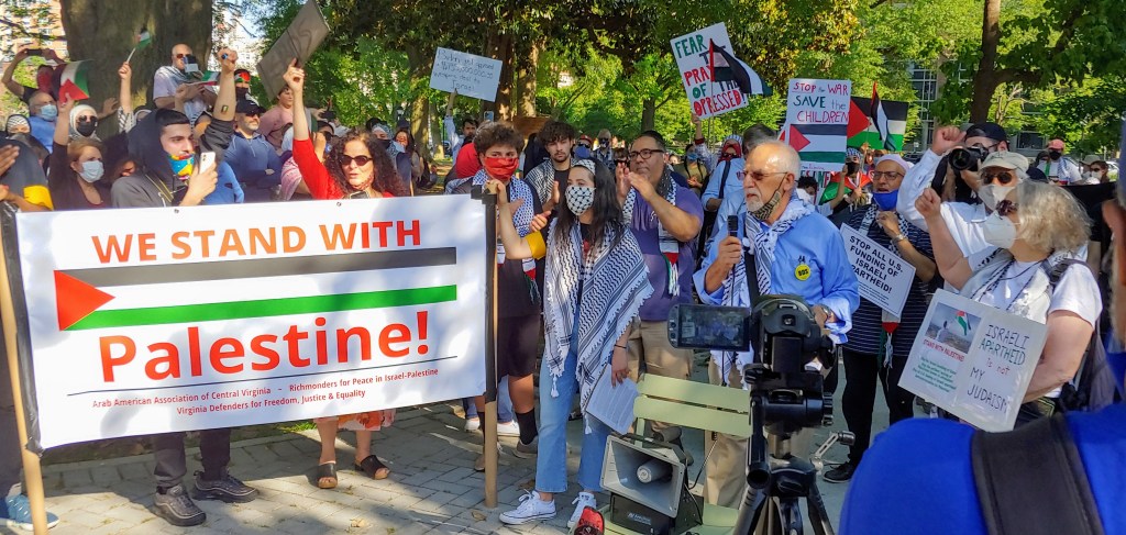 A large crowd at a pro-Palestinian rally behind a banner. Banner reads "We Stand With Palestine!" and lists three organizations: the Arab American Association of Central Virginia, Richmonders for Peace in Israel-Palestine, and Virginia Defenders for Freedom, Justice & Equality. Three speakers, Sanaz Ghodsi, Adeeb Abed, and Nancy Wein are pictured in the foreground. 