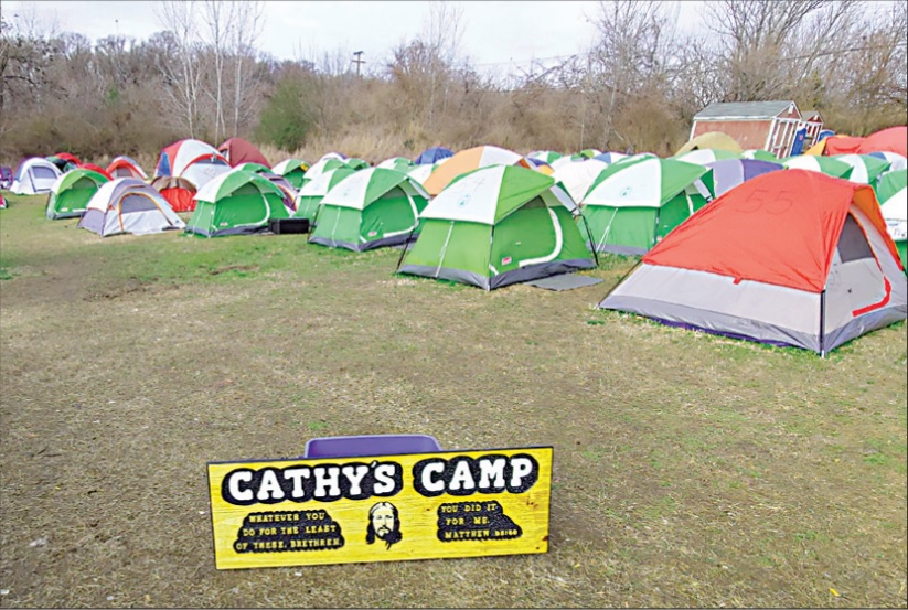 Photograph of rows of dozens of tents in a field under an overcast sky. In the foreground, there is a handmade plywood sign that reads "Cathy's Camp" with a picture of Christ's face on it. 
